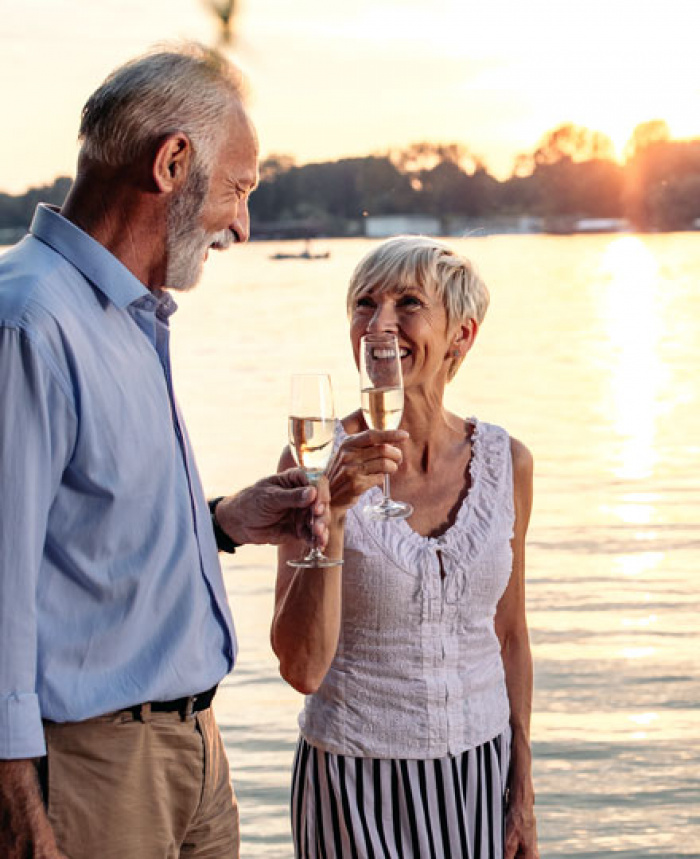 Happy retired couple with champagne glasses walking on beach at sunset